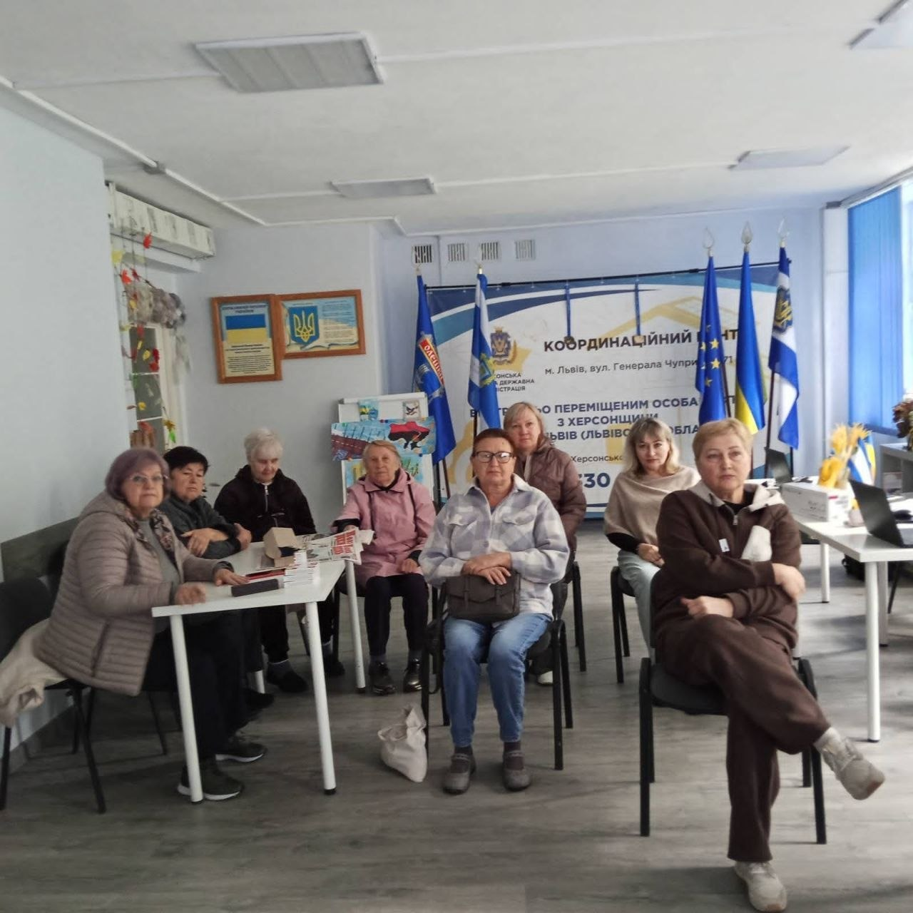 Фотографія, що ілюструє зустріч відвідувачів у Координаційному центрі підтримки цивільного населення «Вільні разом» у Львові, де у світлій кімнаті за білими столами та на стільцях сидить група жінок старшого та середнього віку, які прийшли на освітньо-просвітницькі заходи для переселенців із Херсонщини, при цьому на задньому плані видно великий інформаційний банер центру із зазначенням адреси, п'ять прапорів на підставках — два з яких, включно з прапором Херсонської області, встановлені ліворуч від банера, а три інші, серед яких прапор Європейського Союзу,Державний Прапор України та ще один прапор Херсонської області, розташовані праворуч, тоді як на лівій стіні розміщено патріотичні стенди з гербом та символікою.