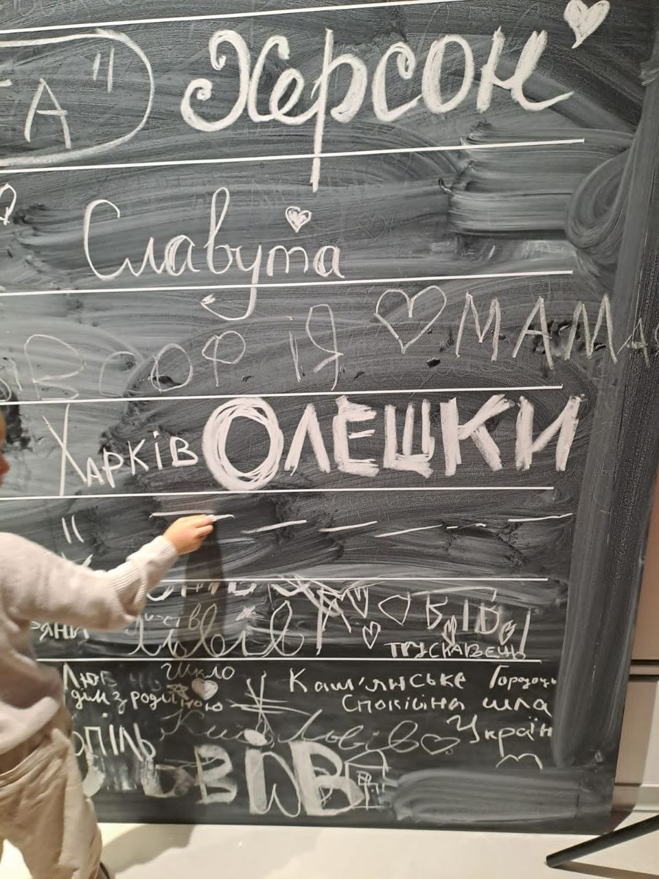 Знімок, на якому дитина ліворуч пише крейдою на великій шкільній дошці, де виведені назви українських міст і селищ, зокрема «Херсон», «Славута», «Харків», «Олешки», «Трускавець» та інші написи з малюнками сердець і будиночків, що ілюструє творчу або інтерактивну частину заходів для родин переселенців із Херсонщини в Координаційному центрі «Вільні разом» у Львові.