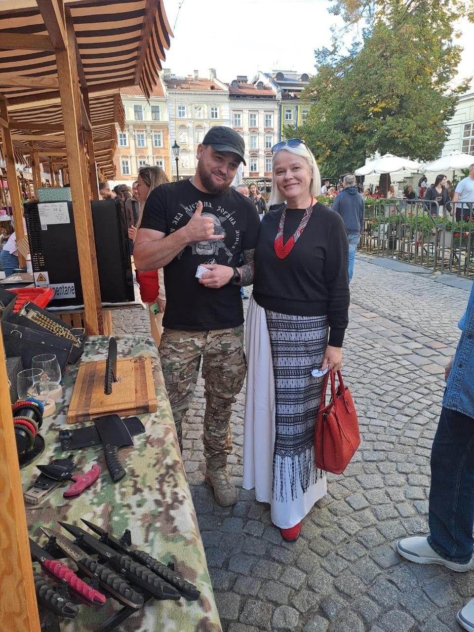 Знімок, на якому на брукованій міській площі біля дерев'яного ярмаркового ятка з виставленими на продаж ножами та сокирами стоять поруч і посміхаються у кадр чоловік у чорній футболці, кепці та камуфляжних штанах, який показує жест «шака», та блондинка в чорному кофті, автентичній довгій спідниці з орнаментом, червоному намисті та з червоною сумкою в руці, що ілюструє дозвілля або прогулянку відвідувачів Координаційного центру «Вільні разом» історичною частиною Львова під час осінніх просвітницьких та культурних заходів.