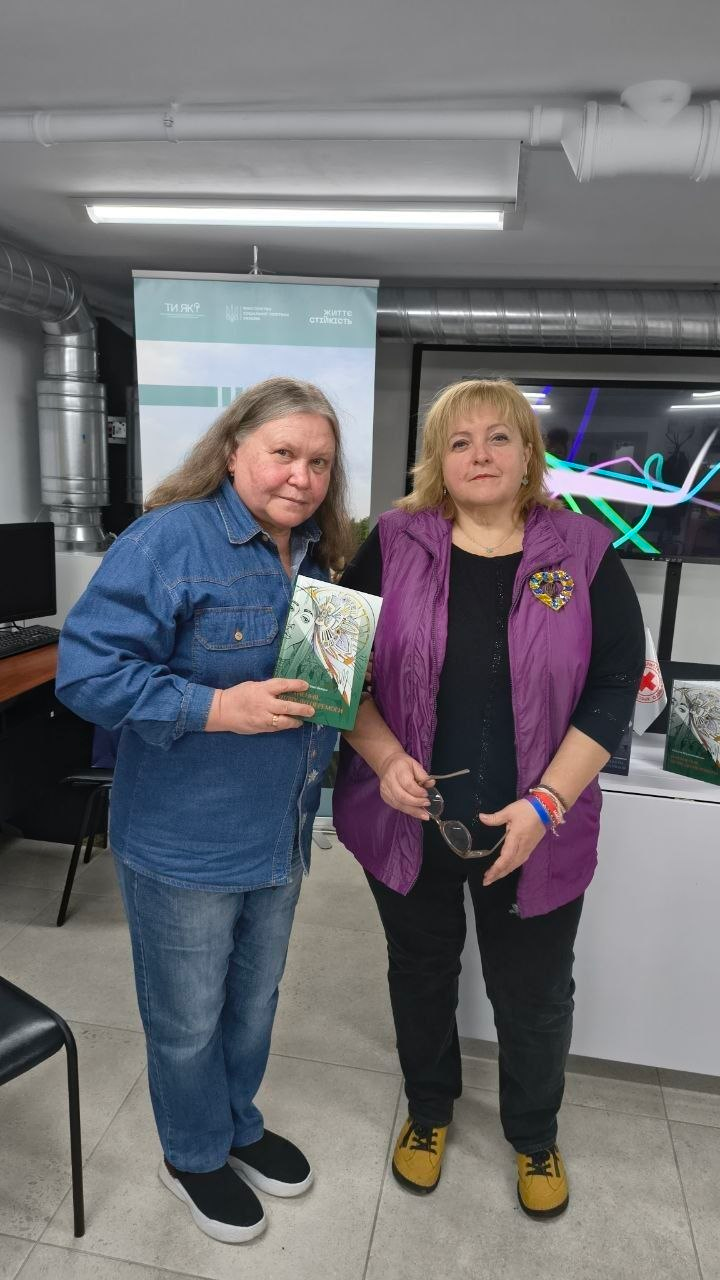 Знімок, на якому у світлому кабінеті Центру життєстійкості у Херсоні стоять поруч і дивляться в кадр дві жінки, де жінка ліворуч з довгим русявим волоссям, одягнена у джинсову сорочку та джинси, тримає в руці книгу в зеленій обкладинці під назвою «Натхнення — шлях до перемоги» авторства Наталі Шушляннікової, а жінка праворуч у фіолетовій безрукавці поверх чорної кофти тримає в руці окуляри, при цьому на задньому плані видно офіційний інформаційний стенд із логотипом програми «Ти як?», великий монітор із абстрактним зображенням, комп'ютерну техніку та елементи вентиляційної системи будівлі.