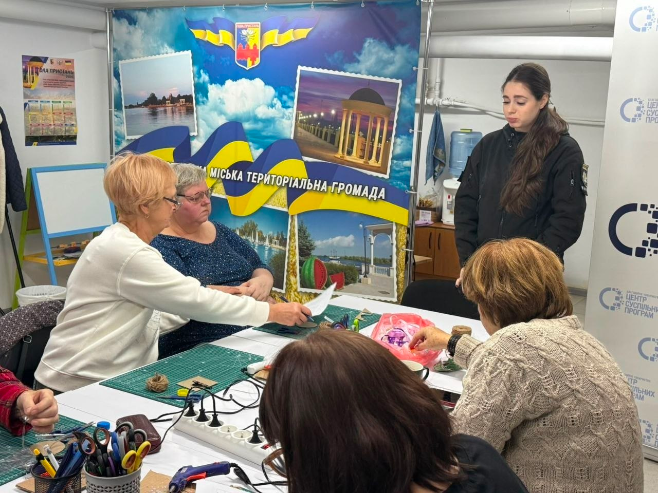Знімок, що ілюструє творчий процес під час арт-терапевтичного майстер-класу в Центрі життєстійкості у Херсоні, де група жінок сидить за великим столом, обладнаним для рукоділля зеленими килимками, ножицями, клеєм та стрічками, і виготовляє патріотичні вироби, при цьому ліворуч жінка у білій кофті передає аркуш паперу іншій учасниці, праворуч за процесом спостерігає дівчина-інструкторка у чорній форменій куртці, а на задньому плані розміщено великий інформаційний банер Голопристанської міської територіальної громади із зображенням набережної, альтанки та герба міста.