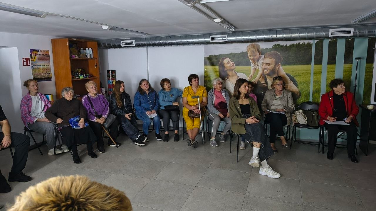 Знімок, що ілюструє тематичну зустріч із психосоціальної підтримки для мешканців області в Центрі життєстійкості у Херсоні, де група жінок різного віку сидить на стільцях півколом уздовж стіни у просторому кабінеті та зосереджено слухає спікера чи виконує завдання з використанням метафоричних асоціативних карт, при цьому праворуч на задньому плані видно велике фотошпалерне зображення щасливої родини на зеленому полі, ліворуч стоїть висока дерев'яна шафа з поличками, на стіні висить інформаційний плакат, а деякі учасниці тримають у руках папери для записів.