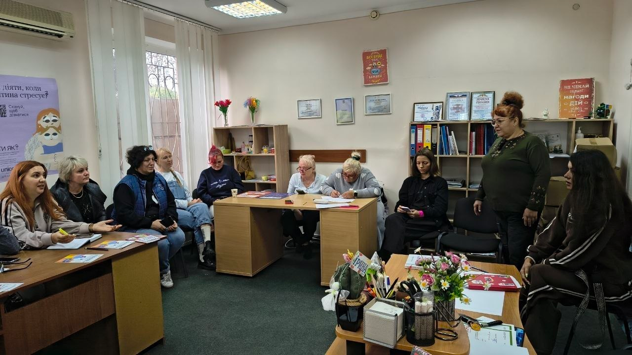 Знімок, що ілюструє проведення інформаційної сесії щодо протидії домашньому насильству та алгоритмів дій у кризових ситуаціях у Центрі життєстійкості у Херсоні, де в кабінеті за робочими столами та на стільцях сидить група жінок, які зосереджено слухають лекторку у темно-зеленій кофті з зібраним у пучок волоссям, яка стоїть праворуч біля стелажа з папками та книгами, при цьому на першому плані ліворуч дівчина з рудим волоссям робить нотатки в зошиті та спілкується з іншими учасницями, у центрі кімнати за столом жінка заповнює бланки документів, а на світлих стінах приміщення розміщено інформаційні плакати програми «Ти як?», сертифікати у рамці, квіти у вазах та гасла підтримки.