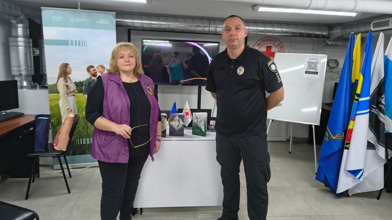 Знімок, на якому у світлому кабінеті Центру життєстійкості у Херсоні поруч стоять і дивляться в кадр жінка у фіолетовій безрукавці поверх чорної кофти з окулярами в руці та чоловік у чорній поліцейській формі з шевронами, які брали участь у заходах із психосоціальної підтримки для мешканців області, при цьому між ними на задньому плані розташований білий стіл із виставленими книгами та маленькими прапорцями України і Червоного Хреста, а в інтер'єрі приміщення видно офіційний мобільний стенд програми «Ти як?», великий монітор, білу магнітну дошку з QR-кодом та кілька великих прапорів на підставках праворуч.