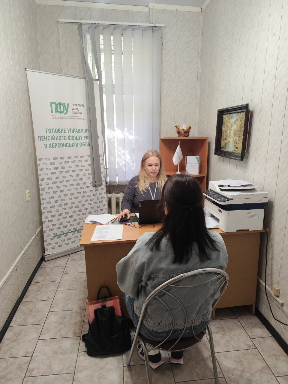 Знімок, що ілюструє прийом переселенців із Херсонщини фахівцем Головного управління Пенсійного фонду України в Херсонській області, який проходить у кабінеті Координаційного центру «Вільні разом» в Одесі за підтримки БФ «Сапорт Херсон», де за дерев'яним кутовим столом навпроти відвідувачки, яка сидить спиною до камери у джинсовій куртці на стільці з металевою спинкою, розташована  жінка-спеціалістка у сірій кофті, яка консультує громадянку та працює за відкритим чорним ноутбуком, при цьому ліворуч у кімнаті встановлено великий вертикальний білий банер-стенд із зеленим логотипом «ПФУ ПЕНСІЙНИЙ ФОНД УКРАЇНИ» та написом «ГОЛОВНЕ УПРАВЛІННЯ ПЕНСІЙНОГО ФОНДУ УКРАЇНИ В ХЕРСОНСЬКІЙ ОБЛАСТІ», а на задньому плані біля вікна з вертикальними жалюзі розміщено невелику дерев'яну шафу з папками та настільним прапорцем, тумбу з великим білим багатофункціональним пристроєм-принтером, картину в темній рамці на стіні та чорний рюкзак відвідувачки на підлозі.