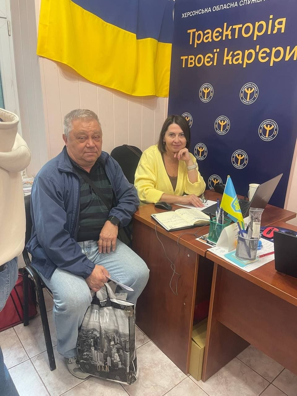 Знімок, що ілюструє проведення консультацій щодо працевлаштування для переселенців із Херсонщини кар'єрним радником Херсонського обласного центру зайнятості, які проходять у Координаційному центрі «Вільні разом» в Одесі за підтримки БФ «Сапорт Херсон», де за дерев'яним офіційним столом поруч із ноутбуком, відкритим блокнотом-органайзером, канцелярією та маленьким настільним прапорцем України сидить жінка-консультантка у світло-жовтій кофті, яка посміхається в кадр, а ліворуч від неї на стільці розмістився відвідувач — сивочолий чоловік у синій куртці, смугастій футболці та блакитних джинсах, який тримає в руках інформаційний буклет та пакет із чорно-білим міським принтом, при цьому на задньому плані встановлено великий темно-синій брендований банер Державної служби зайнятості із жовтим написом «Траєкторія твоєї кар'єри» та круглими логотипами установи, а зверху на стіні розгорнуто великий державний прапор України.