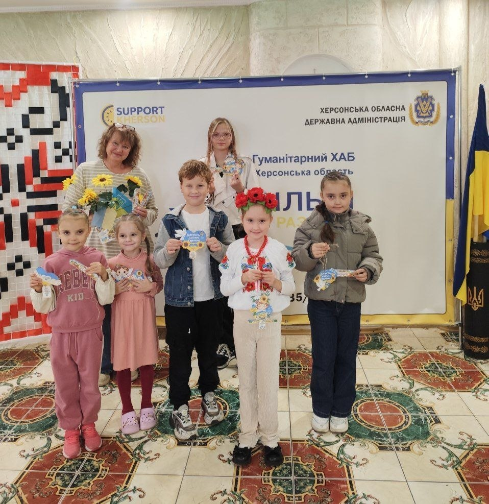 Знімок, що ілюструє завершення творчого майстер-класу в центрі розвитку дітей Херсонщини «Малюк» при Координаційному центрі «Вільні разом» в Одесі за підтримки БФ «Сапорт Херсон», де на тлі великого світлого банера з написами «ХЕРСОНСЬКА ОБЛАСНА ДЕРЖАВНА АДМІНІСТРАЦІЯ», «Гуманітарний ХАБ Херсонська область» та логотипом фонду стоїть група з п'яти дітей різного віку, дівчинки-підлітка та жінки-організаторки ліворуч, які тримають у руках виготовлені власноруч патріотичні вироби та сувеніри з синьо-жовтими елементами, при цьому дівчинка в центрі одягнена у вишиванку та віночок із червоними квітами, хлопчик поруч тримає паперове серце, а жінка ліворуч тримає букет із соняшників, тоді як у кутках кабінету з візерунчастим кахлем на підлозі видно вертикальне панно з червоно-чорним українським орнаментом вишивки ліворуч та державний прапор України праворуч.