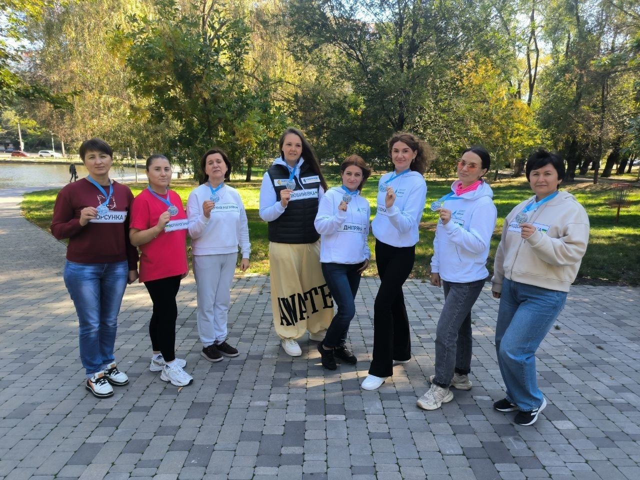 Знімок, що ілюструє участь переселенок із Херсонщини у III благодійному забігу «Об'єднані заради добра» на підтримку українських міст за сприяння Координаційного центру «Вільні разом» в Одесі, БФ «Сапорт Херсон» та БФ «Місто тисячі джерел», де на брукованій алеї міського парку на тлі зелених дерев і водойми стоїть у шеренгу група з восьми усміхнених жінок, які тримають у руках та демонструють на камеру отримані пам'ятні медалі на блакитних стрічках, при цьому на білих худі та спортивних кофтах учасниць закріплені стартові номери з назвами рідних населених пунктів або громад, зокрема «ХЕРСОНКА», «ЛЮБИМІВКА», «ДНІПРЯНИ» та «ТЯГИНКА».