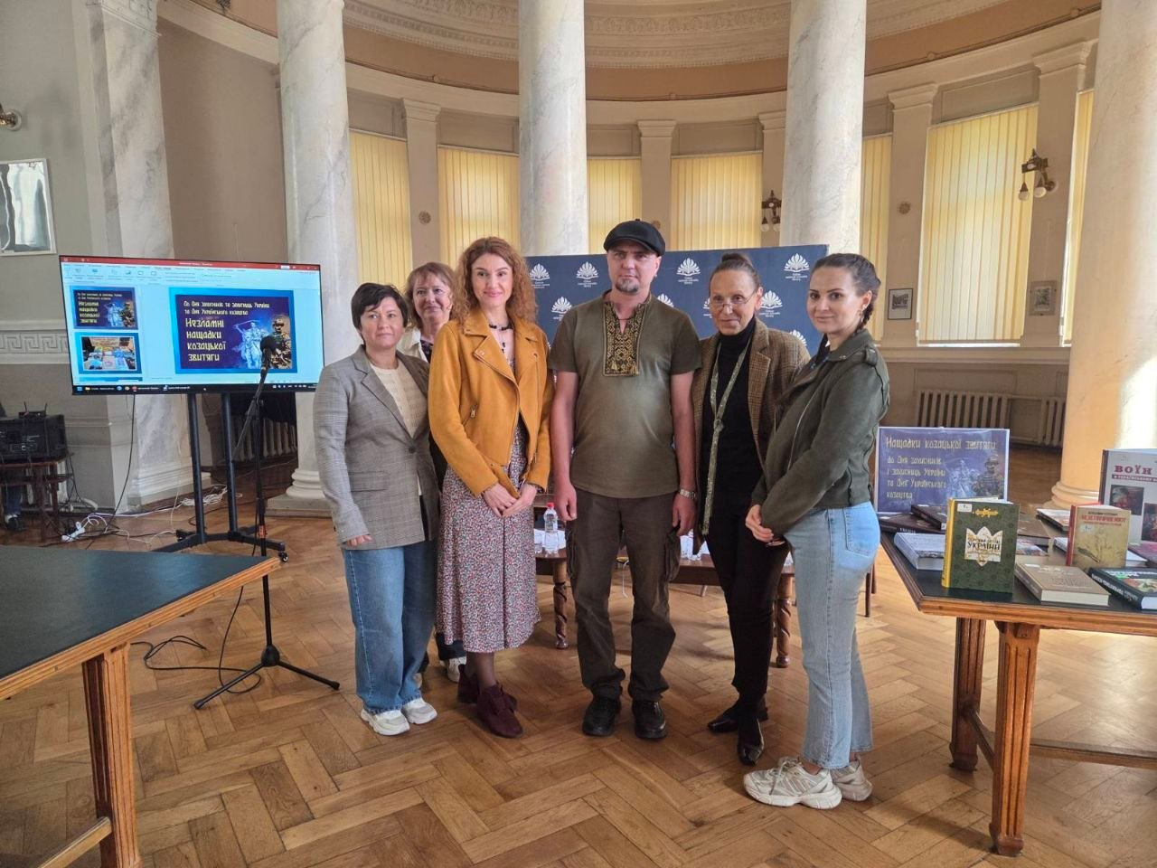 Знімок, що ілюструє відвідування переселенцями з Херсонщини презентації книжково-ілюстративної виставки «Незламні нащадки козацької звитяги» в Одеській національній науковій бібліотеці за сприяння Координаційного центру «Вільні разом» в Одесі та БФ «Сапорт Херсон», де у просторій величній залі з високими світлими колонами, паркетною підлогою та великими вікнами стоїть у шеренгу група організаторів та учасниць заходу, серед яких у центрі чоловік у темно-зеленій футболці з вишивкою та чорній кепці, при цьому ліворуч на стійці встановлено великий телевізійний екран із відкритим слайдом презентації про козацьку звитягу, а праворуч розташовано дерев'яний стіл із виставленими тематичними книгами, інформаційним плакатом та емблемою бібліотеки у рамці.