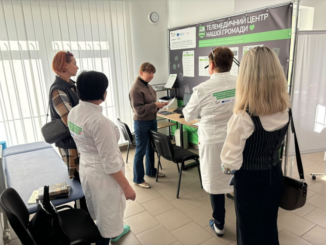 Знімок, що ілюструє ознайомлення учасників заходу з роботою місцевої амбулаторії загальної практики — сімейної медицини під час Дня спільнодії Всеукраїнської програми ментального здоров’я «Ти як?» у Калинівській громаді, де у світлому медичному кабінеті біля великого темного стенда з написом «ТЕЛЕМЕДИЧНИЙ ЦЕНТР НАШОЇ ГРОМАДИ» та логотипом програми стоїть група фахівців і координаторів, зокрема у центрі біля столу з принтером перебуває директорка Департаменту здоров’я Херсонської ОДА Тетяна Карчевич у коричневому кардигані та синіх джинсах, яка тримає в руках аркуш паперу та спілкується з колегами, при цьому на першому плані праворуч спиною до камери стоять жінка-медичний працівник у білому халаті з написом на спині та регіональна координаторка програми Ірина Задорожня у чорній жилетці й блузці, ліворуч біля кушетки розташовані дві інші учасниці зустрічі, а на задньому плані кімнату освітлює велике вікно з вертикальними жалюзі.