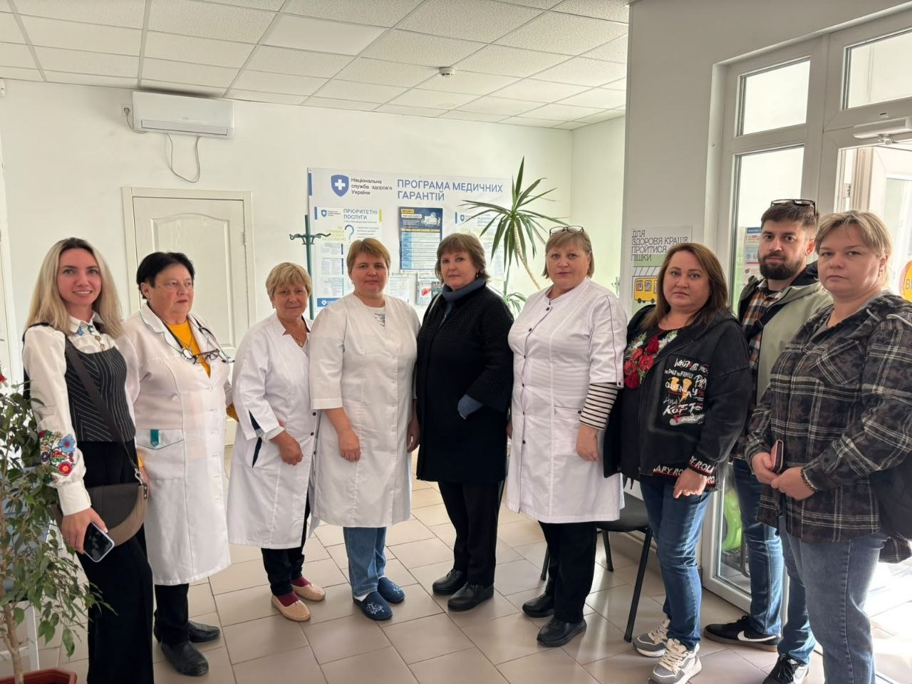 Знімок, що ілюструє відвідування місцевої амбулаторії загальної практики — сімейної медицини під час Дня спільнодії Всеукраїнської програми ментального здоров’я «Ти як?» у Калинівській громаді, на якому група учасників заходу та медичних працівників стоїть разом у світлому холі установи та дивиться в кадр, при цьому крайня ліворуч стоїть регіональна координаторка програми Ірина Задорожня у чорній жилетці й блузці з вишитими рукавами, поруч із нею розташовані три жінки-медики у білих халатах, далі по центру стоять дві учасниці у темному одязі та медичний працівник, а праворуч перебувають жінка в чорній худі з принтом, чоловік у сорочці в клітинку та жінка в кардигані, тоді як на задньому плані видно великий інформаційний стенд Національної служби здоров'я України «Програма медичних гарантій», кімнатну пальму в горщику, кондиціонер на стіні та скляні вхідні двері праворуч.