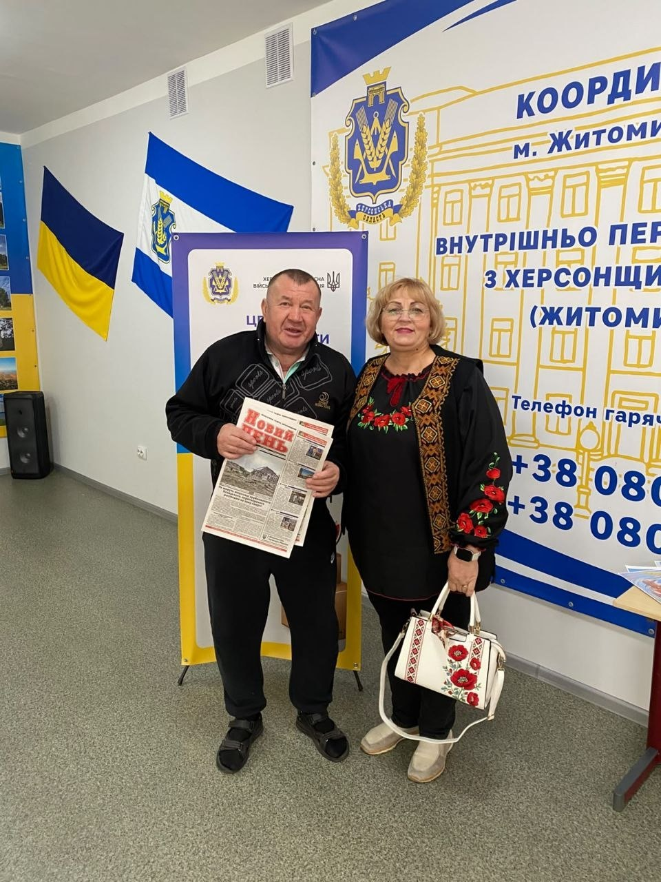 Фотоілюстрація до допису про підтримку переселенців із Херсонщини в житомирському Координаційному центрі «Вільні разом», де на світлині в приміщенні хабу стоять усміхнені чоловік та жінка старшого віку, причому чоловік тримає в руках свіжий випуск обласної газети Херсонщини «Новий день», жінка вбрана в чорну сорочку з вишивкою та тримає білу сумку з квітковим візерунком, а на задньому плані видно великі синьо-жовті інформаційні стенди з гербом Херсонщини та номерами гарячої лінії, тоді як ліворуч на стіні розгорнуто державний прапор України та прапор міста Херсон, що загалом підкреслює турботу про ВПО у Житомирі та забезпечення їх важливою інформацією через друковані видання.