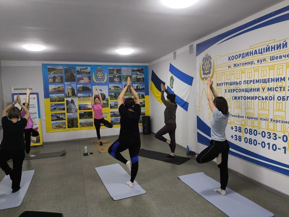 Фотоілюстрація до допису про підтримку переселенців із Херсонщини в житомирському Координаційному центрі «Вільні разом», де на світлині у світлому приміщенні хабу тренерка Наталія Бутинець проводить заняття з йоги для дорослих, під час якого п'ятеро учасниць у спортивному одязі стоять на спеціальних килимках і синхронно виконують вправу на баланс — «позу дерева» зі зведеними над головою долонями, а на задньому плані інтер'єру видно великі інформаційні стенди з гербом і визначними місцями Херсонської області, номерами гарячої лінії, а також закріплені на стіні державний прапор України та прапор міста Херсон.