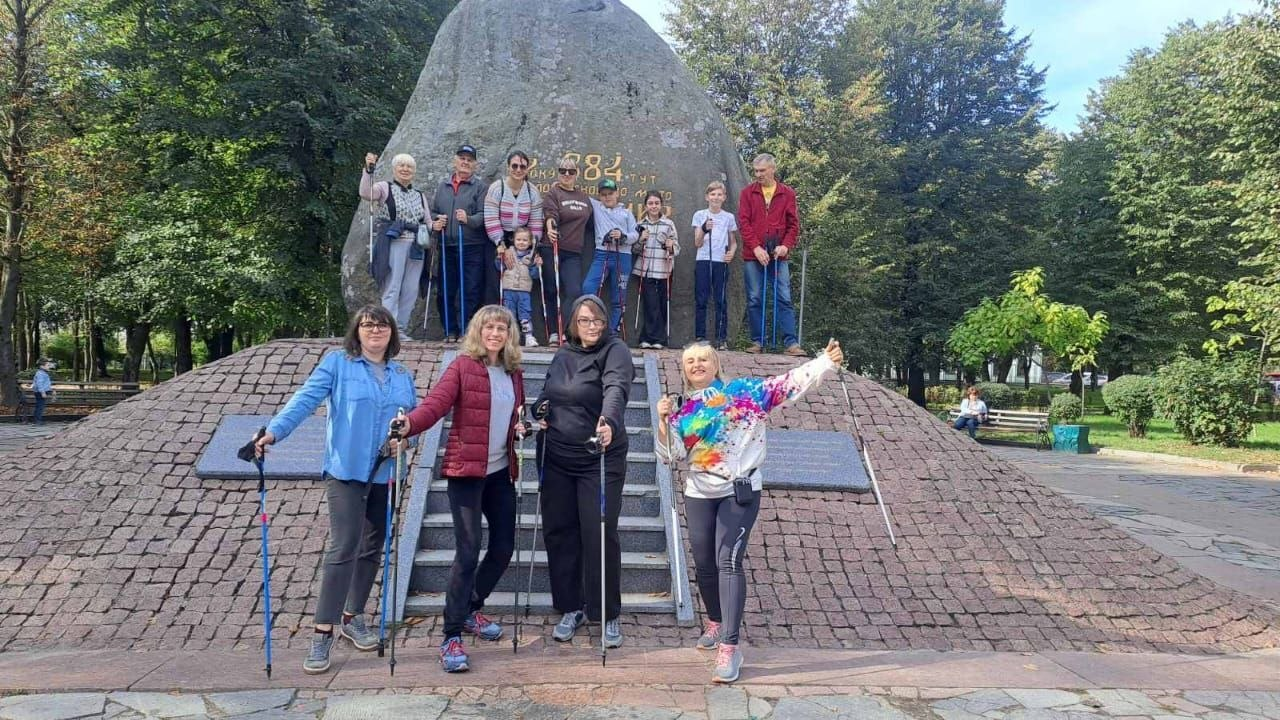Фотоілюстрація до допису про підтримку переселенців із Херсонщини в житомирському Координаційному центрі «Вільні разом», де на світлині група учасників різного віку, серед яких є дорослі та діти, позитивно позує зі спеціальними палицями для скандинавської ходьби на тлі великого пам'ятного каменя Замкової гори в Житомирі, причому частина людей стоїть на узвишші біля підніжжя монумента, а кілька жінок усміхаються попереду на сходах та вимощеному бруківкою майданчику в оточенні зелених дерев міського парку, що візуально демонструє активний відпочинок, оздоровлення та інтеграцію ВПО в нове місто.
