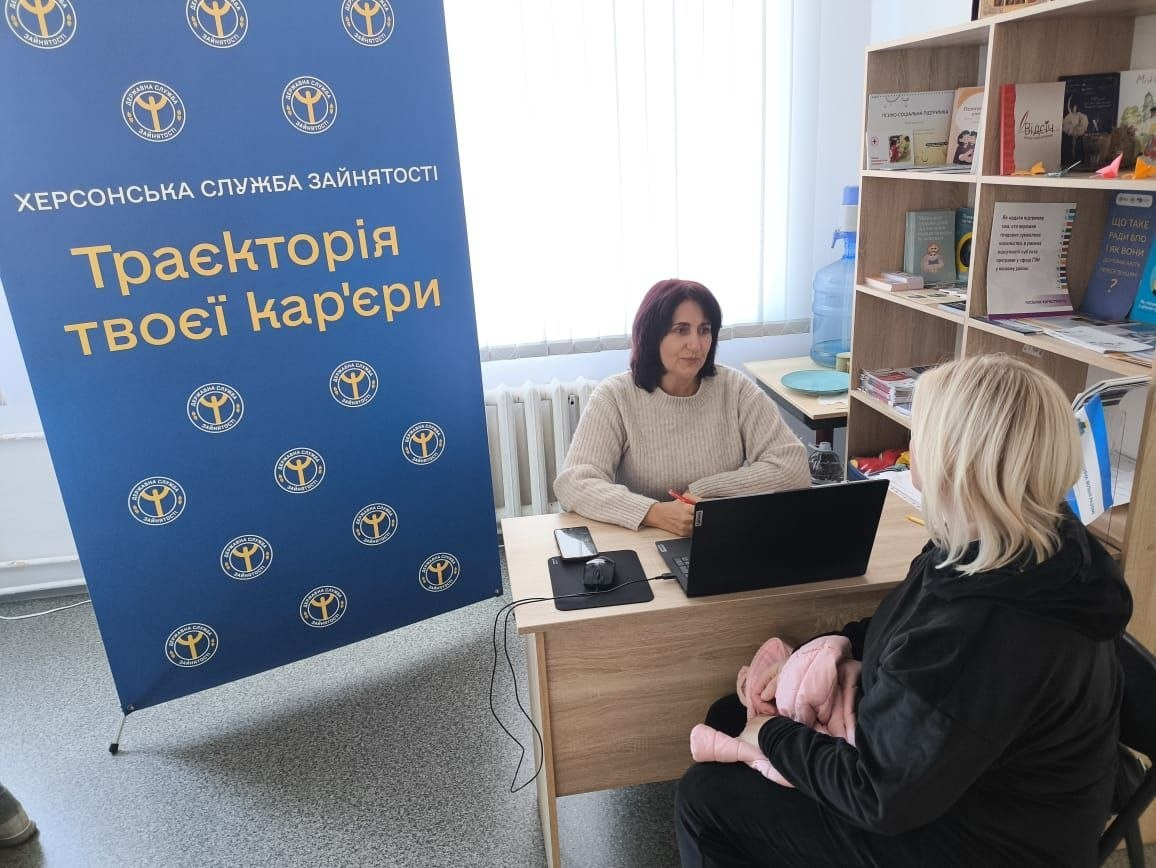 Фотоілюстрація до допису про підтримку переселенців із Херсонщини в житомирському Координаційному центрі «Вільні разом», де на світлині в робочому кабінеті за дерев'яним столом сидить фахівчиня Бериславської філії Херсонського обласного центру зайнятості перед відкритим чорним ноутбуком і консультує жінку-відвідувачку, яка сидить навпроти спиною до об'єктива, а ліворуч розташовано великий синій банер Державної служби зайнятості з написами «ХЕРСОНСЬКА СЛУЖБА ЗАЙНЯТОСТІ» та «Траєкторія твоєї кар'єри», тоді як праворуч на задньому плані видно офісний стелаж із книгами та інформаційними брошурами.