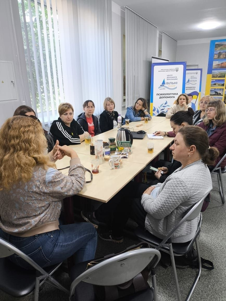Фотоілюстрація до допису про підтримку переселенців із Херсонщини в житомирському Координаційному центрі «Вільні разом», де на світлині в офісному приміщенні хабу психологиня ГО «Авенір» Олена Ратинська проводить чергову зустріч групи підтримки «Коло жінок», під час якої учасниці сидять навколо великого спільного столу з напоями, уважно слухають та обговорюють важливі теми, а на задньому плані кімнати встановлено мобільні банери центру «Вільні разом» із написами про психологічну та юридичну допомогу переселенцям.