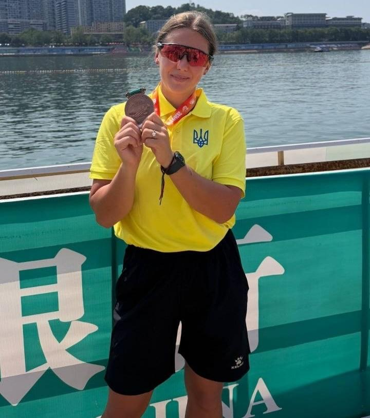Українська спортсменка Олена Скворцова у жовтій футболці-поло з синім тризубом на грудях та чорних шортах стоїть на понтоні на тлі річки в Ханчжоу (Китай). Вона посміхається, носить спортивні окуляри з червоними лінзами і тримає в руках медаль, яку здобула на міжнародних змаганнях з веслування на човнах «Дракон». На задньому плані — широка водойма та міський пейзаж протилежного берега під ясним небом.