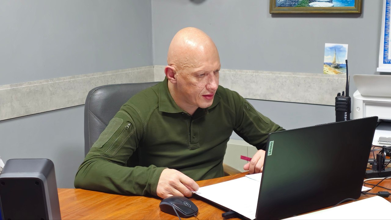 Заступник начальника Херсонської ОВА Євгеній Ігнатенко сидить за дерев'яним столом у робочому кабінеті перед відкритим чорним ноутбуком під час засідання обласної комісії з питань надання матеріальної допомоги мешканцям області. Він одягнений у тактичну сорочку кольору хакі з довгими рукавами, уважно дивиться на екран комп'ютера та тримає руку на документах. На столі поруч із ноутбуком розташована комп'ютерна миша, праворуч на задньому плані стоїть рація, а на сірій стіні позаду висить картина у рамці.