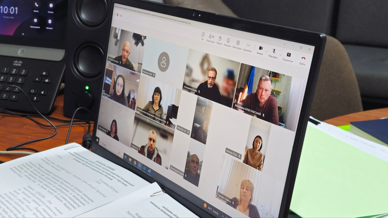 Екран ноутбука, на якому відкрито вікно онлайн-відеоконференції Microsoft Teams під час засідання обласної комісії щодо виплат матеріальної допомоги жителям Херсонщини. На екрані відображаються вікна учасників онлайн-зустрічі: у лівому верхньому кутку присутній заступник начальника Херсонської ОВА Євгеній Ігнатенко, у верхньому ряду поруч — заступник директора Департаменту з питань цивільного захисту та оборонної роботи Херсонської ОДА Володимир Бєляєв, у другому ряду ліворуч — заступник директора Департаменту соціального розвитку Херсонської ОДА Олена Масюткіна, в цьому ж ряду крайня праворуч — начальник управління взаємодії з правоохоронними органами Херсонської ОДА Юлія Іванчук. Ноутбук стоїть на робочому столі, поруч лежать паперові документи з порядком денним засідання, а ліворуч на задньому плані видно стаціонарний офісний телефон та аудіоколонку.