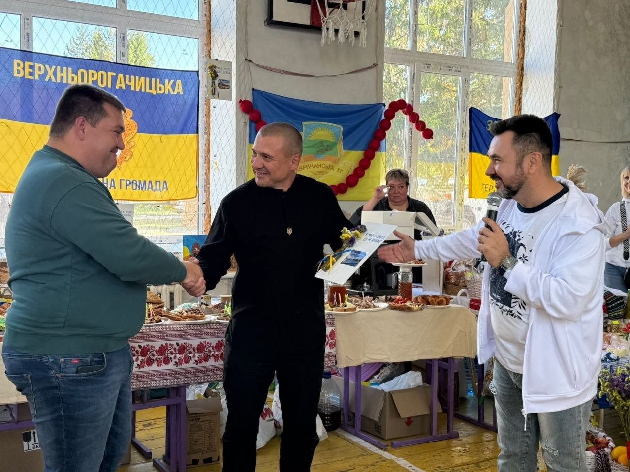 Начальник Верхньорогачицької СВА Артур Давидов (по центру, у чорній сорочці з маленьким золотим тризубом) посміхається і тисне руку представнику Окнянської громади Одеської області (ліворуч, у зеленому светрі та синіх джинсах). Одночасно з цим заступник директора Департаменту реалізації гуманітарної політики Херсонської ОДА Андрій Ярьоменко (праворуч, у білій кофті) говорить у мікрофон та передає їм нагородний диплом за посіте друге місце у конкурсі кулінарних традицій на заході «Смакуємо у рідному селі». Нагородження проходить у спортивному залі на тлі ярмаркових столів із випічкою та медом, а на стіні позаду учасників висять прапори, зокрема банер «Верхньорогачицька селищна територіальна громада».
