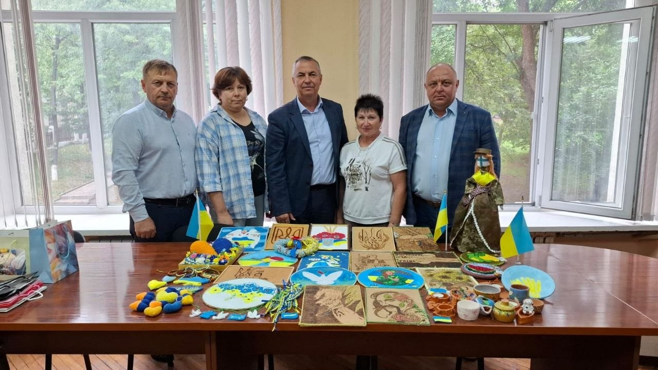 Голова Окнянської громади Подільського району Одеської області Олег Бєлоус (посередині, у темно-синьому піджаку та блакитній сорочці) разом із представниками Верхньорогачицької та Окнянської громад позує в кабінеті за довгим столом, на якому презентовано творчі роботи жителів громад для благодійного ярмарку «Смакуємо у рідному селі», де на столі розкладено різноманітні патріотичні вироби ручної роботи: малюнки та картини, в'язані синьо-жовті сувеніри, декоративні тарілки з гербом і картою України, глиняний посуд, а також велика сувенірна лялька-мотанка в традиційному вбранні праворуч, причому композицію прикрашають маленькі державні прапорці, а всі п'ятеро учасників дивляться в кадр на тлі великих вікон, за якими видно зелені дерева.