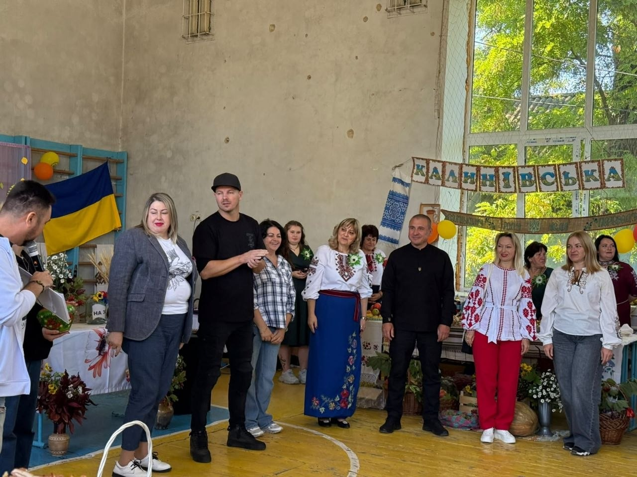 Директор Департаменту реалізації гуманітарної політики Херсонської ОДА Віолета Царькова (у картатій сорочці та джинсах), заступник директора Департаменту Андрій Ярьоменко (крайній ліворуч, з мікрофоном у руці) та начальник Верхньорогачицької СВА Артур Давидов (у чорній сорочці та штанах) разом із представниками та учасниками громад позують у спортивному залі під час благодійного заходу «Смакуємо у рідному селі» в межах проєкту «Пліч-о-пліч: згуртовані громади», де перед ними виступає чоловік у чорній футболці та кепці, поруч стоїть жінка в сірому піджаку, а на задньому плані праворуч облаштовано виставкову локацію із рушниками та написом «Калинівська», де інші учасниці в традиційних українських вишиванках підтримують захід аплодисментами.
