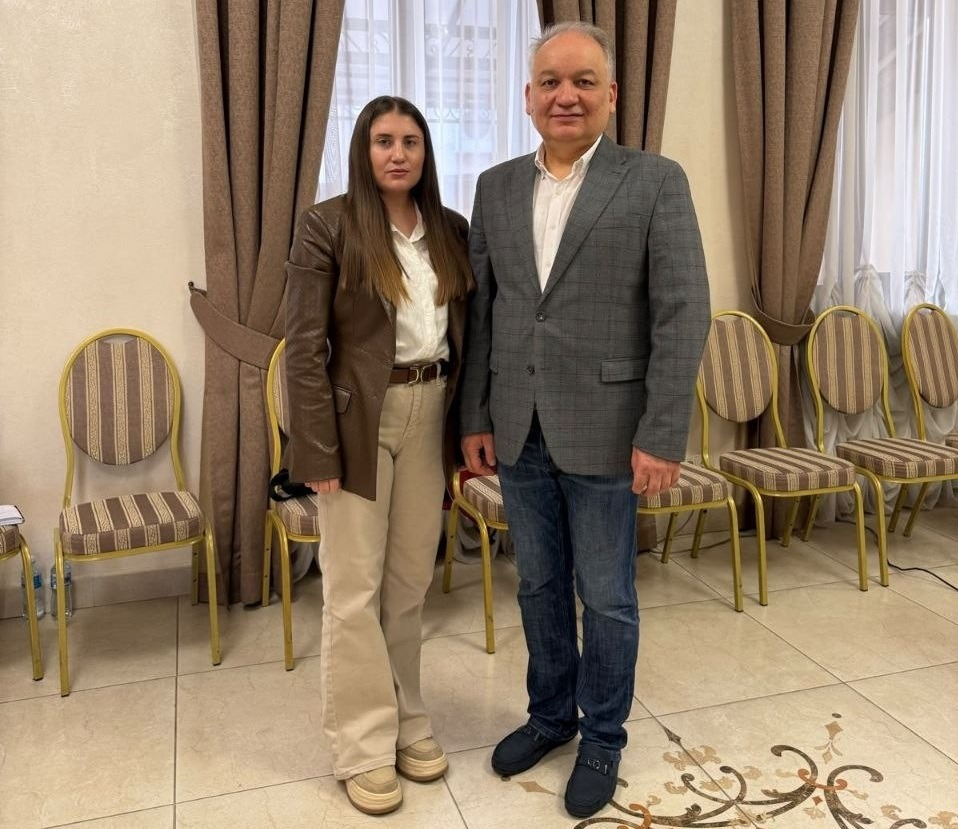Начальниця служби у справах дітей Верхньорогачицької селищної ради (ліворуч, у коричневому шкіряному піджаку, білій сорочці та світлих штанах) позує для спільного фото з чоловіком у сірому класичному піджаку, білій сорочці та синіх джинсах у залі засідань під час дводенного експертного семінару «Документування порушень прав дітей в умовах війни» в Одесі, де на задньому плані видно інтер'єр приміщення зі світлими шторами, великими коричневими портьєрами та вишикуваними в ряд м'якими стільцями із золотистим каркасом.
