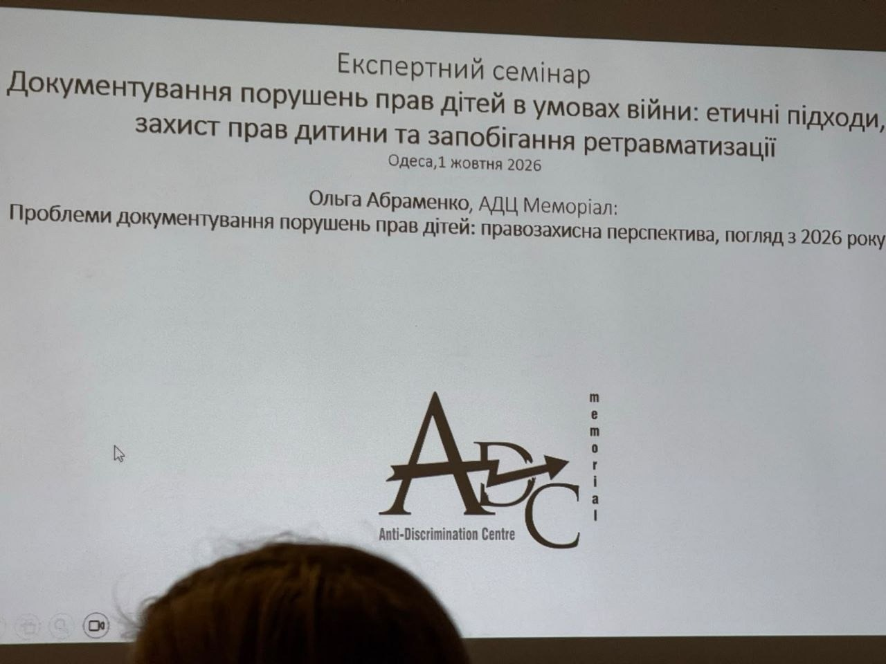 Титульний слайд презентації на великому білому екрані під час дводенного експертного семінару в Одесі, де чітко видно заголовок заходу «Документування порушень прав дітей в умовах війни: етичні підходи, захист прав дитини та запобігання ретравматизації», дату проведення «Одеса, 1 жовтня 2026», тему доповіді «Проблеми документування порушень прав дітей: правозахисна перспектива, погляд з 2026 року» від експертки Ольги Абраменко з АДЦ «Меморіал», а в нижній частині слайда по центру розміщено офіційний темно-сірий логотип організації з написами Anti-Discrimination Centre та Memorial, причому на передньому плані в самому низу кадру помітно темне волосся одного з учасників заходу, який слухає лекцію.