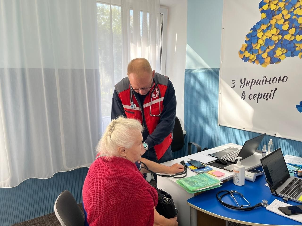 Медичний працівник мобільної бригади Товариства Червоного Хреста України у фірмовому червоно-синьому жилеті з емблемою організації та стетоскопом на шиї вимірює артеріальний тиск літній жінці-переселенці з Херсонщини в координаційному центрі «Вільні разом» у Хмельницькому, де пацієнтка у яскраво-рожевій кофті сидить на стільці біля робочого столу медика, на якому розкладені медичні тонометри, документи, антисептик, стоять два відкриті ноутбуки, а на синьо-білій стіні праворуч висить великий плакат із патріотичним малюнком карти України з жовто-блакитних сердець та патріотичним гаслом «З Україною в серці!».