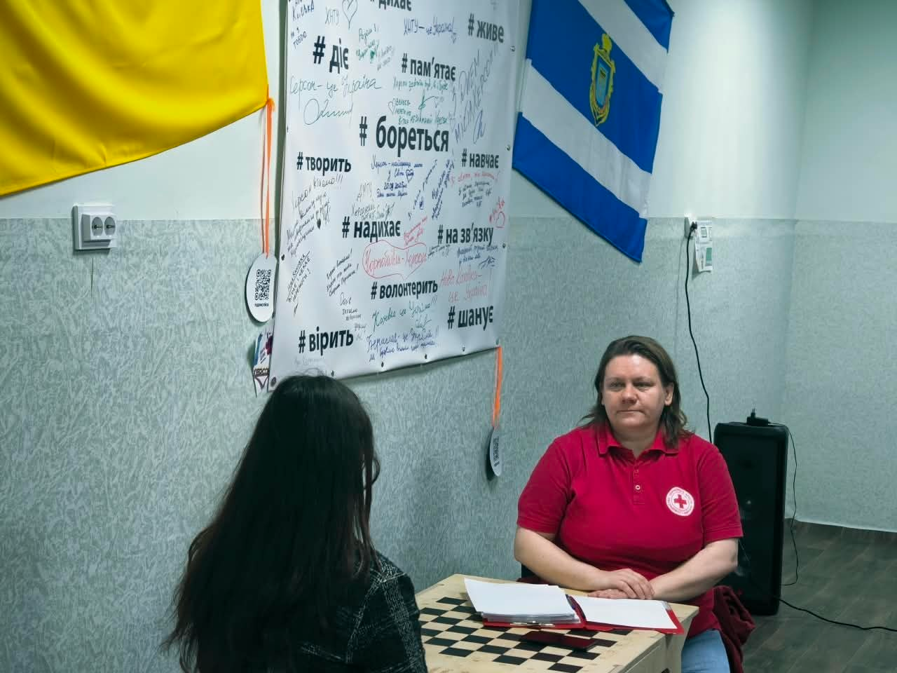 Психологиня Товариства Червоного Хреста України у фірмовій червоній футболці-поло з білим круглим логотипом організації на грудях проводить індивідуальну консультацію для переселенки з Херсонщини в координаційному центрі «Вільні разом» у Хмельницькому. Психологиня сидить за столом, стиснувши руки перед собою, і уважно слухає жінку із довгим темним волоссям, яка сидить спиною до камери на передньому плані ліворуч. На столі перед ними лежать робочі документи та папери. Інтер'єр кабінету прикрашений патріотичною символікою: на стіні ліворуч висить державний прапор України, по центру розташований великий білий плакат із написами від руки та чорними мотиваційними хештегами, серед яких «#бореться», «#діє», «#вірить», «#волонтерить», «#пам'ятає», а праворуч на стіні закріплено синьо-білий прапор міста Херсон із міським гербом.