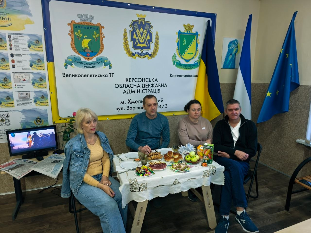 Команда координаційного центру «Вільні разом» у Хмельницькому (дві жінки та двоє чоловіків) сидить за гостинним столом із частуваннями, фруктами, солодощами та соком під час презентації простору, причому на стіні позаду них встановлено великий офіційний білий стенд із гербами та написами «Великолепетиська ТГ», «Херсонська обласна державна адміністрація, м. Хмельницький, вул. Зарічанська 14/3» та «Костянтинівська ТГ», а праворуч стоять Державний прапор України, прапор Херсонської області та прапор Європейського Союзу, водночас ліворуч на столі поруч із ними розміщено ввімкнений комп'ютерний монітор із патріотичною заставкою та свіжі випуски газет, а на крайній лівій стіні закріплено інформаційний плакат із фотокартками діяльності хабу.