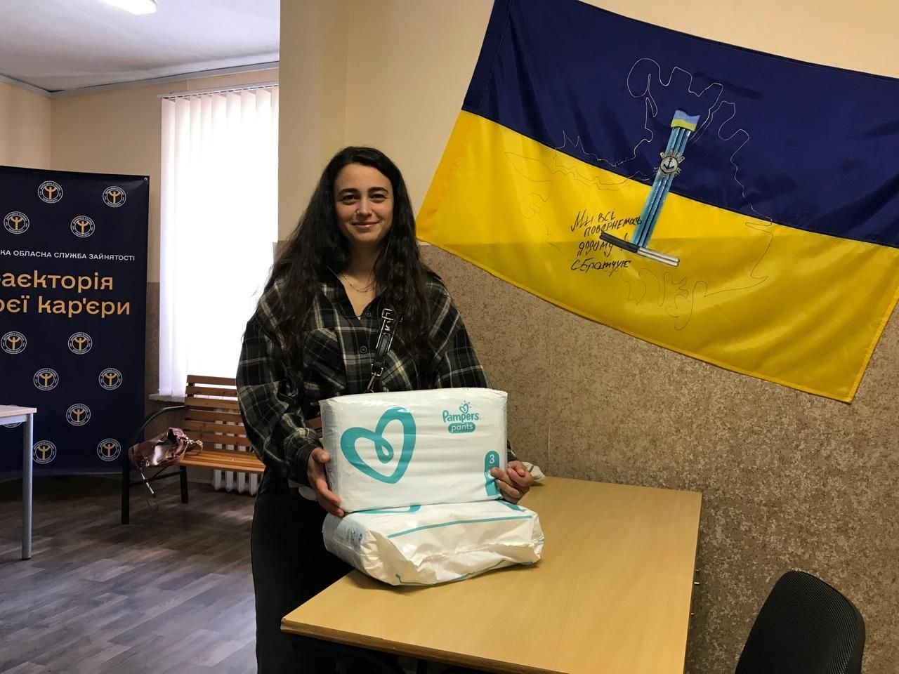 Молода жінка-переселенка з Херсонщини посміхається і тримає в руках великі упаковки дитячих підгузків Pampers pants, які вона отримала в межах системної підтримки в координаційному центрі «Вільні разом» у Хмельницькому, причому вона стоїть біля дерев'яного офісного столу в приміщенні хабу, де на стіні праворуч висить Державний прапор України з нанесеними контурами карти Херсонської області, символічним малюнком херсонського малого герба по центру та патріотичним написом від руки «Ми всі повернемося додому, С. Братчук», а ліворуч на задньому плані встановлено темно-синій брендований стенд Державної служби зайнятості з гаслом «Траєкторія твоєї кар'єри».