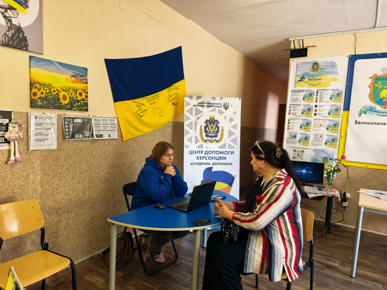 Юристка координаційного центру у фірмовій синій кофті з капюшоном сидить за напівкруглим синім столом перед відкритим ноутбуком і надає індивідуальну юридичну консультацію переселенці з Херсонщини в координаційному центрі «Вільні разом» у Хмельницькому, де поруч із ними встановлено білий інформаційний ролап-стенд із написом «Херсонська обласна державна адміністрація. Центр допомоги херсонцям. Юридична допомога», а на стіні позаду висить великий Державний прапор України з написами від руки, ліворуч від якого розміщено яскраву картину з соняшниковим полем, а праворуч на задньому плані видно великі стенди з символікою хабу та гербом Великолепетиської громади.