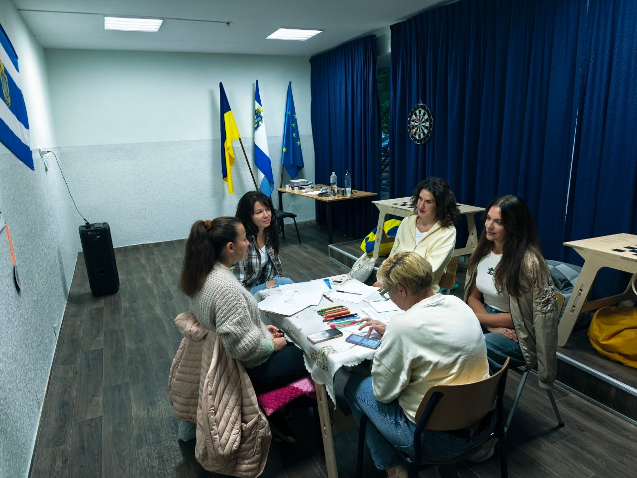 Четверо жінок-переселенок із Херсонщини отримують фахову допомогу психолога під час зустрічі групи підтримки в координаційному центрі «Вільні разом» у Хмельницькому, де вони разом із консультанткою сидять навколо прямокутного столу, застеленого світлою скатертиною, на якій розкладені папери, блокноти та кольорові олівці для арттерапії, причому на стіні ліворуч висить прапор міста Херсона, а на задньому плані стоять Державний прапор України, прапор Херсонської області та прапор Європейського Союзу. Великі вікна праворуч повністю завішені щільними темно-синіми портьєрами, біля яких облаштовано зону з дерев'яними шаховими столами.