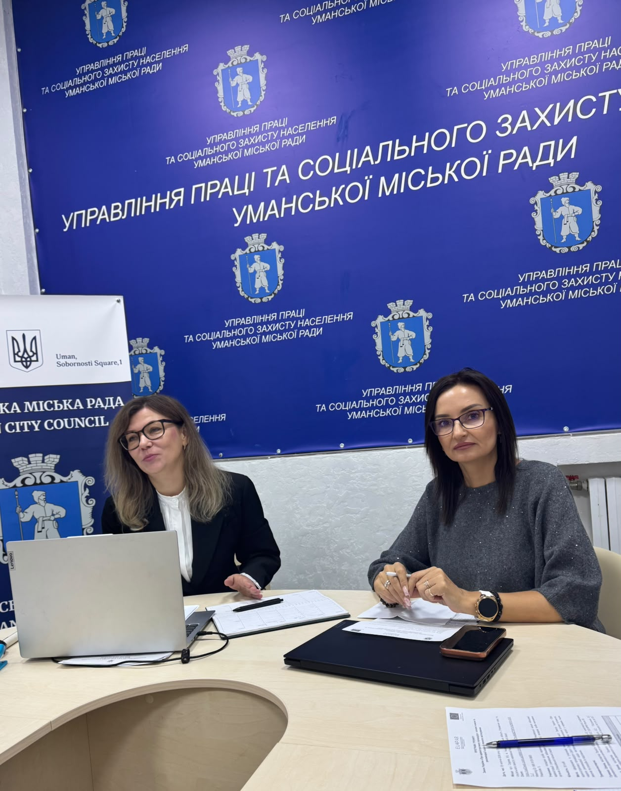 Начальниця Олешківської МВА Тетяна Гасаненко (праворуч, в окулярах та темній кофті) разом із колегою (ліворуч, у піджаку та білій блузці) сидить за робочим столом під час практичного семінару щодо застосування Закону України «Про адміністративну процедуру» в Умані, причому перед ними розкладено паперові роздруківки, ручки, блокноти, мобільні телефони та відкритий ноутбук, за якими вони опрацьовують алгоритми ведення адміністративних справ у межах проєкту Європейського Союзу EU4PAR III, а на задньому плані встановлено великий брендований синій банер із гербами та офіційним білим написом «Управління праці та соціального захисту населення Уманської міської ради» та білим вертикальним банером «Уманська міська рада / Uman City Council» у лівій частині кадру.