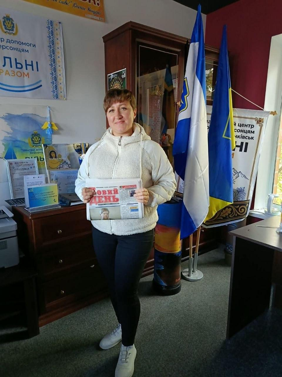 Жінка-переселенка з Херсонщини тримає в руках свіжий примірник друкованої газети «Новий день» під час видачі преси в координаційному центрі «Вільні разом» в Івано-Франківську, де вона одягнена у світлу теплу кофту з капюшоном, чорні штани та білі кросівки і позує в кабінеті хабу на тлі Державного прапора України, прапора Херсонської області, розставлених ліворуч на столі офіційних подяк і грамот установи, а також білого банера з написом «Центр допомоги херсонцям «Вільні разом»».