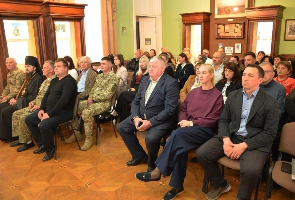 Представники координаційного центру «Вільні разом» у Кропивницькому та ВПО з Херсонщини беруть участь у Молитовному сніданку, де у вишуканій залі з класичним дерев'яним паркетом та світло-зеленими стінами на стільцях сидить велика кількість запрошених гостей, серед яких на першому плані праворуч сидить чоловік у темно-сірому піджаку та блакитній сорочці, поруч із ним — жінка у бордовому светрі та темних штанах, ліворуч від неї — чоловік у класичному синьому костюмі, а в лівому ряду присутні військовослужбовці у камуфляжній формі ЗСУ, священнослужитель у чорному облаченні та інші офіційні особи й учасники заходу, які уважно слухають виступи спікерів.