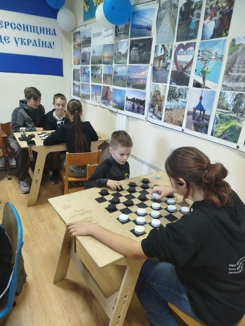 Юні переселенці з Херсонщини грають у настільні ігри під час спортивної зустрічі в координаційному центрі «Вільні разом» у Кропивницькому, де на передньому плані за дерев'яним столом хлопчик та дівчина захоплено грають у шашки, розставляючи чорні та білі фішки на ігровій поверхні, стилізованій під шахівницю, водночас на задньому плані за іншим столом троє підлітків грають у шахи, а сама кімната прикрашена білими та блакитними повітряними кульками, патріотичним банером «Херсонщина — це Україна!» ліворуч та великим настінним фотоколажем зі знімками краєвидів Херсонської області та мирного життя громад.