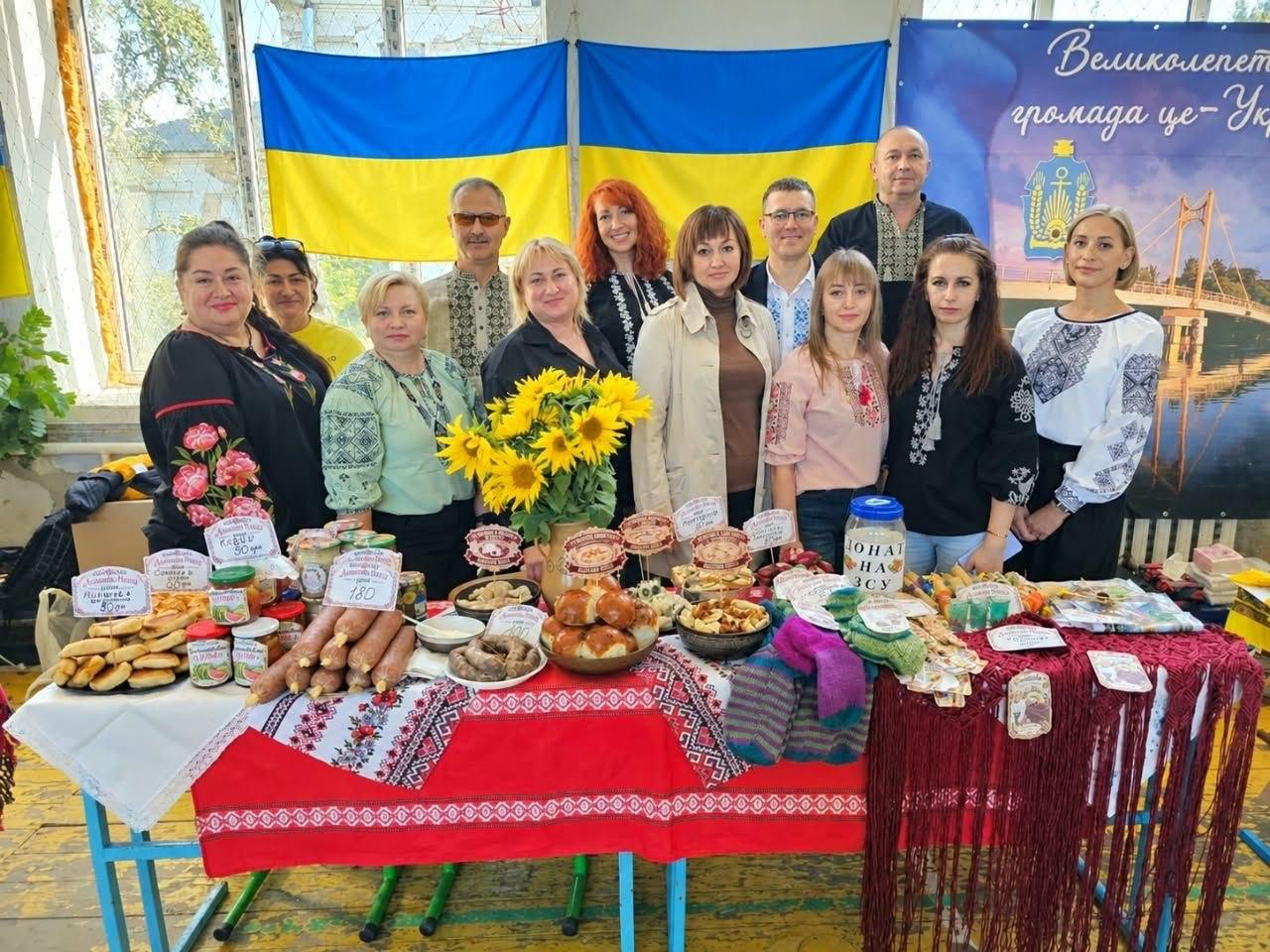 Велика команда координаційного центру «Вільні разом» у Кропивницькому та представники Великолепетиської громади позують для спільного фото за пишним виставковим столом, де вони вибороли перше місце у гастрономічному конкурсі на фестивалі «Смакуємо в рідному селі», причому на передньому плані на великому столі, застеленому вишиваною скатертиною та червоним рушником із торочками, презентовано різноманітні українські кулінарні традиції для конкурсної програми: домашні ковбаси, свіжу випічку, пиріжки, консервацію, пампушки, м'ясні вироби та великий букет яскравих соняшників по центру, а праворуч стоїть банка з написом «Донат на ЗСУ» та розкладено патріотичні в'язані сувеніри, водночас учасники заходу, серед яких багато жінок та чоловіків у традиційних українських вишиванках, радісно посміхаються в кадр у спортивному залі на тлі двох великих синьо-жовтих Державних прапорів України та офіційного банера праворуч із гербом і написом «Великолепетиська громада це — Україна!».