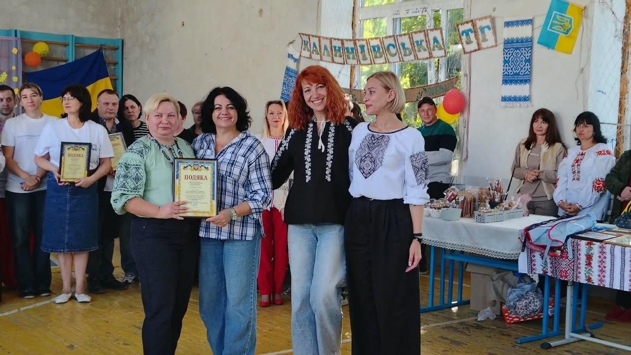 Директор Департаменту реалізації гуманітарної політики Херсонської ОДА Віолета Царькова (друга ліворуч, у картатій сорочці в клітинку та блакитних джинсах) разом із колегами, учасниками та представниками координаційного центру «Вільні разом» у Кропивницькому позує для спільного фото під час благодійного ярмарку на підтримку ЗСУ в межах фестивалю «Смакуємо в рідному селі», де вона спільно з жінкою у світло-зеленій вишиванці тримає в руках отриману офіційну грамоту-подяку у рамці, а по центру поруч із ними стоїть жінка в чорній вишиванці з розпущеним рудим волоссям, праворуч від якої перебуває фахівчиня у білій вишиванці та широких чорних штанах, причому на задньому плані в спортивному залі присутні інші гості заходу, облаштовано ярмаркові столи з випічкою та сувенірами під вивіскою «Калинівська ТГ», а ліворуч на стіні закріплено Державний прапор України, водночас праворуч на стіні висить синьо-білий вишитий рушник та малий Державний Герб України на тлі жовто-блакитної стрічки.
