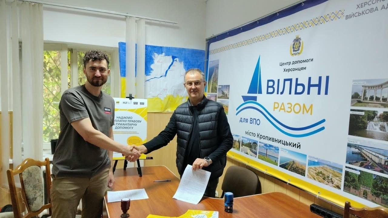 Директор Центру допомоги херсонцям міста Кропивницького «Вільні разом» Іван Дударь (праворуч, у чорній стьобаній жилетці та окулярах) тисне руку представнику мобільної команди БО «100% життя. Кропивницький» (ліворуч, у сірій футболці) над робочим столом у кабінеті хабу під час підписання Меморандуму про співпрацю для підтримки переселенців із Херсонської області, причому керівник центру тримає в лівій руці підписаний примірник офіційної угоди, а на столі перед ними лежать документи, ручка та синя канцелярська печатка, водночас на задньому плані розташований великий білий брендований банер із логотипом у вигляді синього вітрильника, офіційними гербами, фотокартками краєвидів та великими написами «Центр допомоги херсонцям ВІЛЬНІ РАЗОМ для ВПО місто Кропивницький», а на стіні ліворуч висить малюнок карти Херсонщини у синьо-жовтих кольорах та встановлено жовто-білий інформаційний ролап-стенд.
