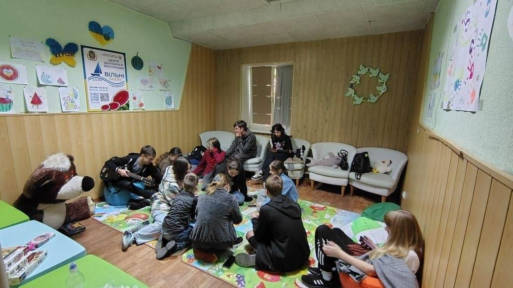 Група підлітків-переселенців із Херсонщини зібралася в ігровій кімнаті координаційного центру «Вільні разом» у Кропивницькому, де частина дітей сидить колом на яскравому дитячому килимку на підлозі та займається приготуванням вафельних тортів зі згущеним молоком під час кулінарного майстер-класу, а інші підлітки розмістилися поруч на м'яких безкаркасних кріслах-мішках і білих диванах біля стіни, оздобленої дерев'яними панелями, причому ліворуч на світло-зеленій стіні розвішані дитячі малюнки з кавунами, патріотичний метелик та офіційний плакат хабу, а на передньому плані зліва сидить велика плюшева іграшка у вигляді ведмедя.