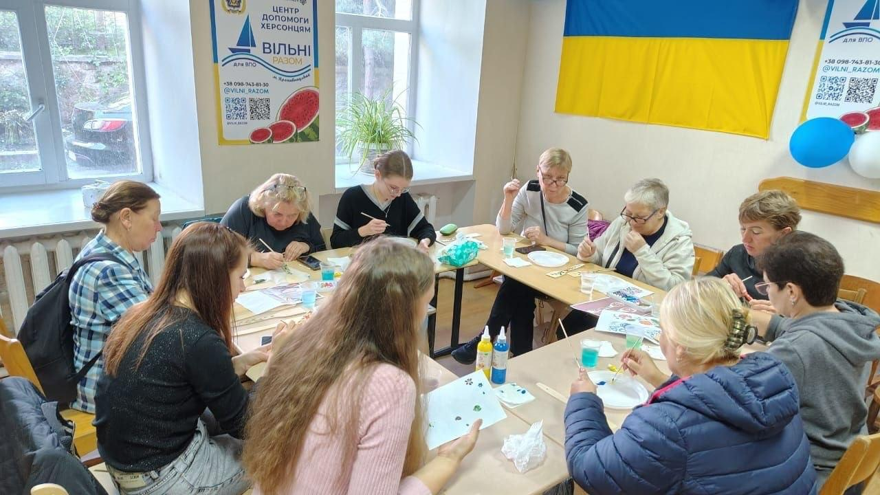 Велика група жінок-переселенок із Херсонщини разом із майстринями бере участь у творчому майстер-класі з розпису дерев’яних ложок, який відбувся напередодні Дня українського козацтва в координаційному центрі «Вільні разом» у Кропивницькому, де тринадцятеро учасниць різного віку сидять за великим П-подібним дерев'яним столом у світлій кімнаті хабу і захоплено розмальовують фарбами сувенірні вироби, використовуючи пензлики, пластикові палітри, стаканчики з водою та тюбики з різнокольоровим акрилом, причому інтер'єр приміщення прикрашений великим Державним прапором України на центральній стіні, синьо-білими повітряними кульками праворуч, а ліворуч біля великого вікна з кімнатною рослиною встановлено офіційний інформаційний плакат із зображенням кавуна та контактами центру допомоги херсонцям.