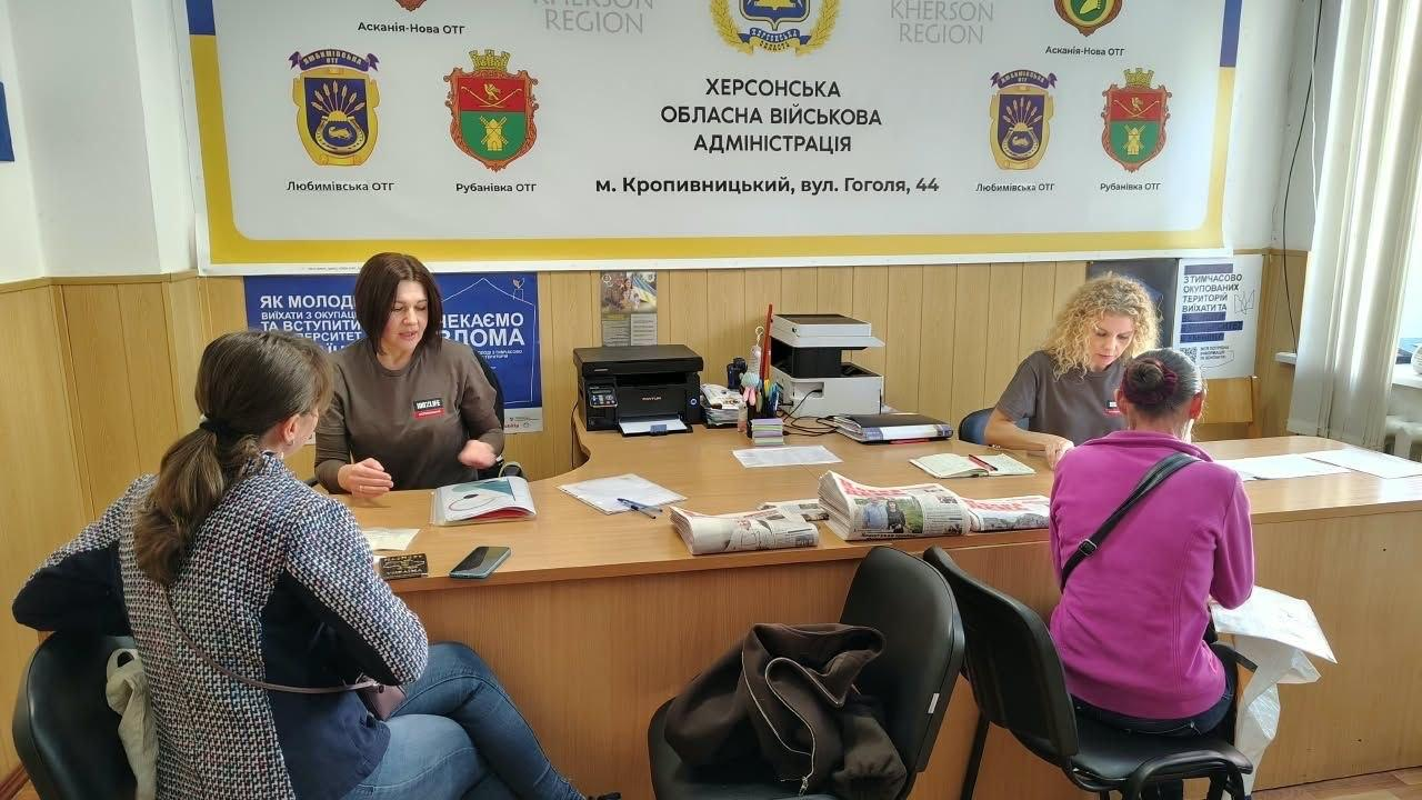 Дві співробітниці координаційного центру «Вільні разом» у Кропивницькому надають консультації та видають свіжі примірники друкованих газет «Новий день» внутрішньо переміщеним особам з Херсонщини, які сидять на стільцях перед великим кутовим робочим столом, де розкладено офіційну документацію, канцелярію, бланки заяв та свіжі випуски преси, причому за столом ліворуч сидітиме консультантка в темно-сірому світшоті, яка спілкується з відвідувачкою у піджаку в дрібну клітинку, а праворуч інша співробітниця з кучерявим світлим волоссям опрацьовує звернення жінки у яскравій рожевій кофті, водночас на задньому плані кабінету встановлено принтер, багатофункціональний пристрій, розвішано інформаційні плакати про вступ до університетів, а на стіні по центру розміщено великий офіційний білий стенд із гербами громад та написами «Херсонська обласна військова адміністрація, м. Кропивницький, «Любимівська ОТГ», «Рубанівська ОТГ» та «Асканія-Нова ОТГ».
