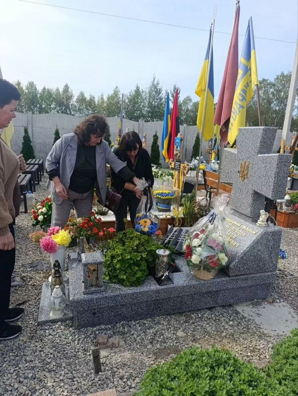 Начальниця Мирненської селищної військової адміністрації (друга ліворуч, у сірому брючному костюмі) разом із рідними загиблого воїна покладає квіти до могили захисника України Івана Лінейцева на кладовищі під час пам'ятного заходу в межах обласного проєкту «Хронологія мужності», де очільниця громади та мати полеглого бійця схилилися над гранітним пам'ятником у формі хреста з викарбуваним ім'ям героя, прикрашаючи його кошиком із жовто-блакитними штучними квітами, поруч із пам'ятником на могилі розставлені свіжі живі квіти, декоративні зелені кущі, лампадки та розгорнута кам'яна книга, праворуч від могили на флагштоках підняті Державні прапори України та стяг військового підрозділу, а на задньому плані за бетонним парканом височіють зелені дерева під світлим небом.