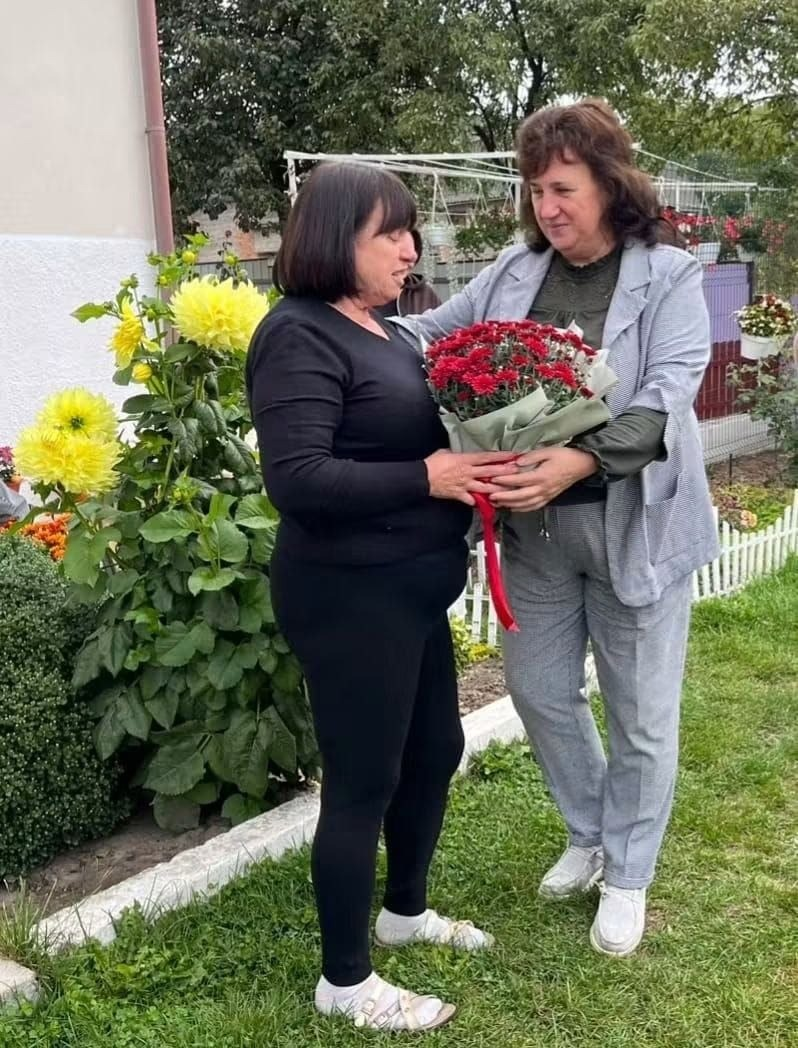 Начальниця Мирненської селищної військової адміністрації (праворуч, у сірому брючному костюмі) обіймає за плече мати загиблого захисника України Івана Лінейцева та передає їй великий букет червоних хризантем під час зустрічі на подвір'ї приватного будинку в межах обласного проєкту «Хронологія мужності», де мати полеглого воїна одягнена в чорну кофту та темні штани і з вдячністю приймає квіти, стоячи на зеленому газоні біля будинку, поруч із яким росте великий кущ із яскраво-жовтими жоржинами та облаштовано декоративний білий парканчик, а на задньому плані видно зелені дерева, металеву садову арку.