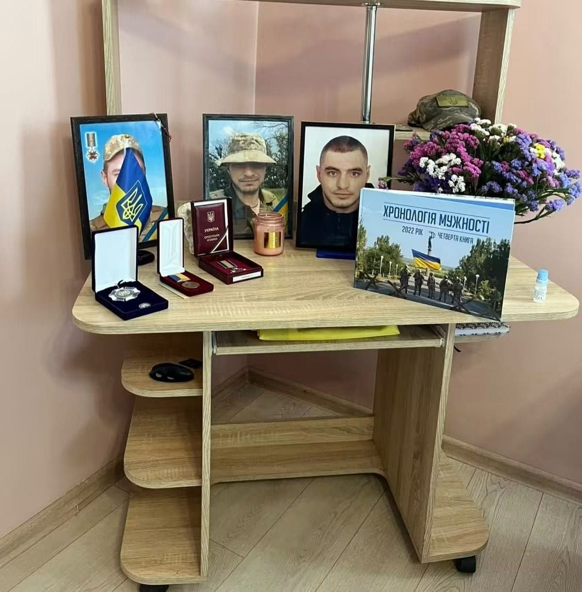 Куточок пам'яті загиблих Героїв, облаштований на дерев'яному комп'ютерному столі в кімнаті, де ліворуч у ряд встановлено три портрети Героїв у військовій формі та цивільному одязі, перед якими розкладено відкриті оксамитові футляри з державними нагородами, орденами та медалями, поруч стоїть запалена ароматична свічка в скляній баночці, по центру на столі розміщено велику пам'ятну книгу «Хронологія мужності. 2022 рік. «Четверта книга»», а праворуч стоїть великий букет із різнокольорових хризантем, над яким на верхній поличці стелажа лежить військова камуфляжна кепка під світло-рожевою стіною.