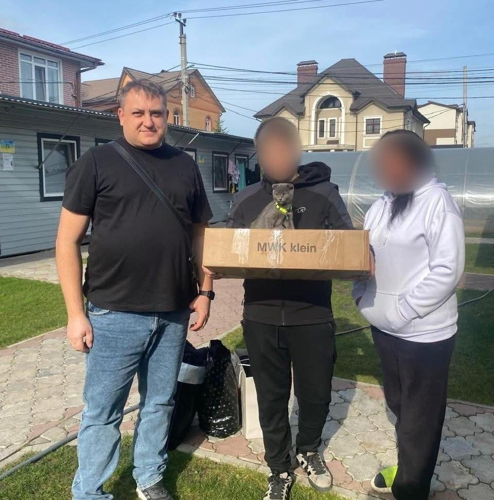 Фахівець служби у справах дітей Чулаківської сільської ради (ліворуч, у чорній футболці та синіх джинсах) стоїть поруч із нещодавно повернутою з тимчасово окупованих територій родиною на подвір'ї будинку, де чоловік по центру тримає в руках велику картонну коробку гуманітарної допомоги з написом «MWK klein», з якої виглядає маленьке сіре кошеня з жовтим нашийником, а праворуч стоїть жінка у білій кофті з капюшоном та темних штанах, обличчя евакуйованих громадян приховано з міркувань безпеки, а самі вони зафіксовані на тлі житлових будинків, зеленого газону та викладеної тротуарною плиткою доріжки під ясним небом.