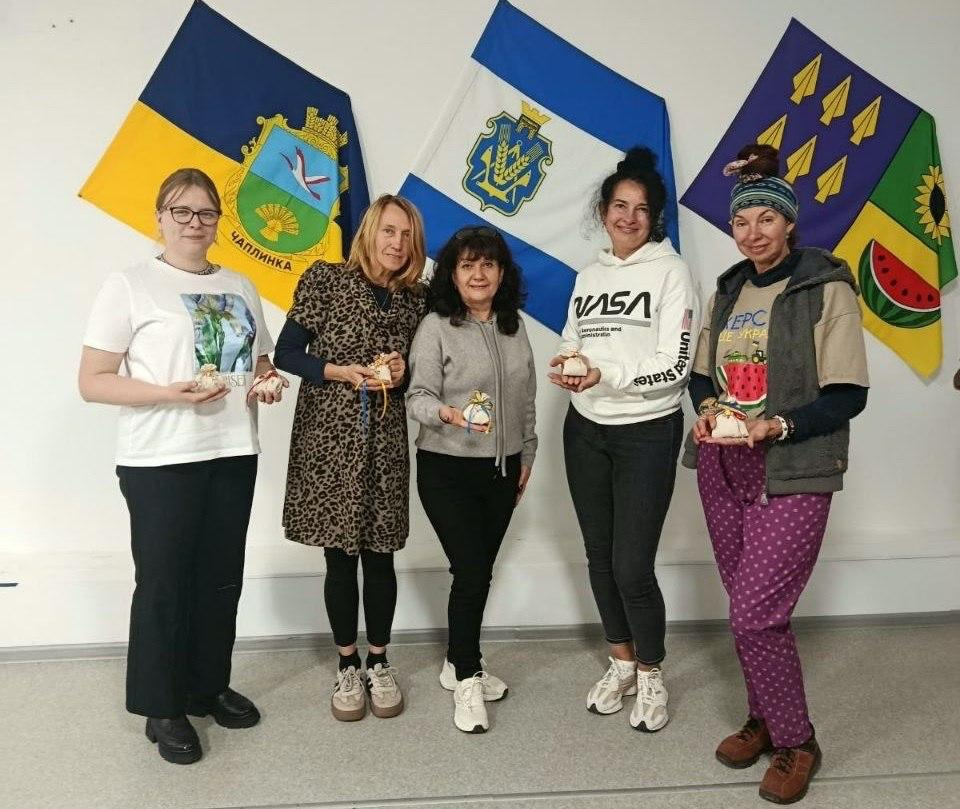П'ять жінок — представниці координаційного центру «Вільні разом» у Черкасах та переселенки з Херсонщини — позують для спільного фото та демонструють виготовлені власноруч сувеніри-обереги у вигляді маленьких мішечків напередодні Дня Захисників і Захисниць України, де вони вишикувалися в один ряд у світлій кімнаті хабу, причому крайня ліворуч одягнена в білу футболку та чорні штани, жінка поруч — у леопардову сукню, по центру стоїть жінка в сірому светрі та чорних штанах, праворуч від неї — жінка у білому худі з написом «NASA» та чорних джинсах, а крайня праворуч — жінка в жилетці, фіолетових штанях у горошок та головній пов'язці, які зафіксовані на тлі чотирьох патріотичних прапорів на білій стіні: Державного прапора України з гербом Чаплинки, прапора Херсонської області, синьо-жовтого прапора Бериславського району та зеленого прапора з малюнком розрізаного кавуна.