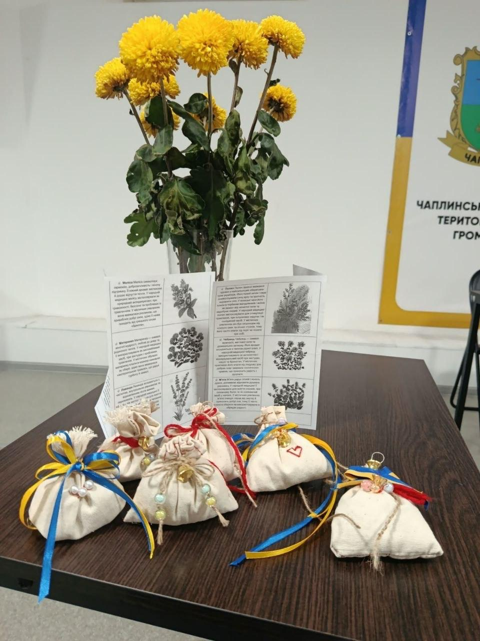 Шість виготовлених власноруч патріотичних сувенірів-оберегів у вигляді маленьких світлих тканинних мішечків, перев'язаних синьо-жовтими та червоно-білими стрічками з намистинами, розкладено на кутку коричневого дерев'яного столу в координаційному центрі «Вільні разом» у Черкасах під час майстерки напередодні Дня Захисників і Захисниць України, де позаду оберегів стоїть відкритий друкований інформаційний буклет із малюнками та описом цілющих рослин, поруч із яким у скляній вазі стоїть великий пишний букет яскраво-жовтих осінніх хризантем, а праворуч на задньому плані на білій стіні частково видно офіційний жовто-білий стенд із написом «Чаплинська територіальна громада» та її гербом.